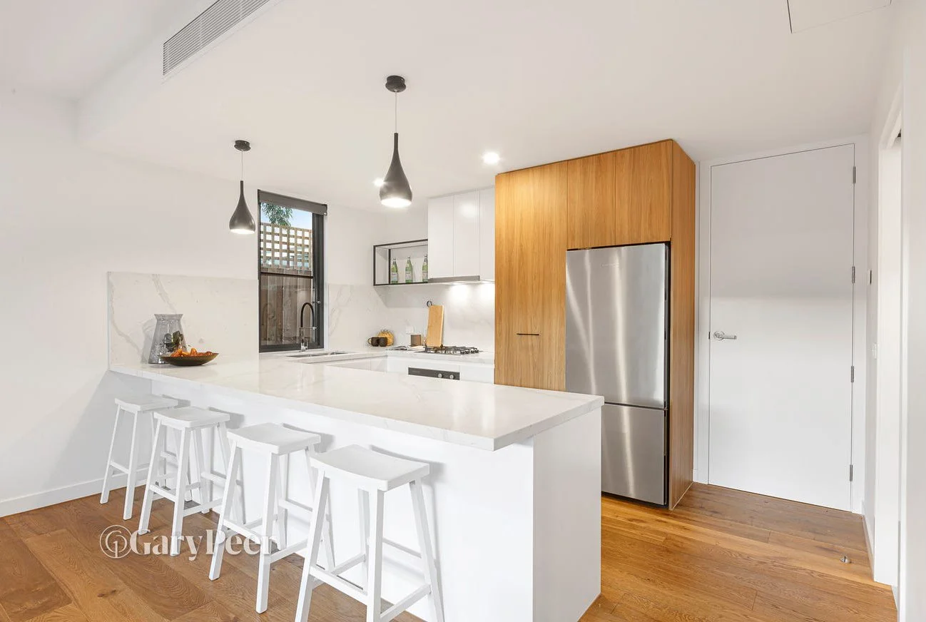 Modern kitchen with white walls and cabinets, wooden flooring, stainless steel refrigerator, black hanging pendant lights, and a small breakfast bar with white stools.