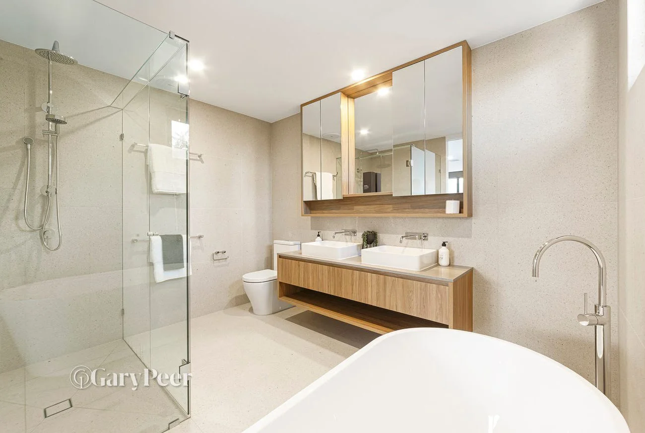 Modern bathroom with a glass-enclosed shower, a freestanding bathtub, a double vanity with vessel sinks, a large mirror, beige tile walls, and minimal decor.
