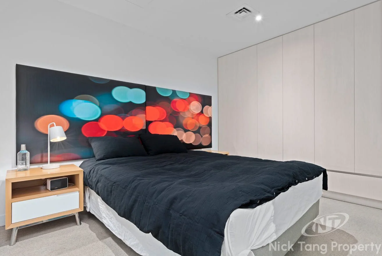 A modern bedroom with a queen-sized bed, black bedding, and pillows, a nightstand with a lamp and water bottle, a digital alarm clock, a large wall art with colorful bokeh lights, white closet doors, and a ceiling with recessed lighting.