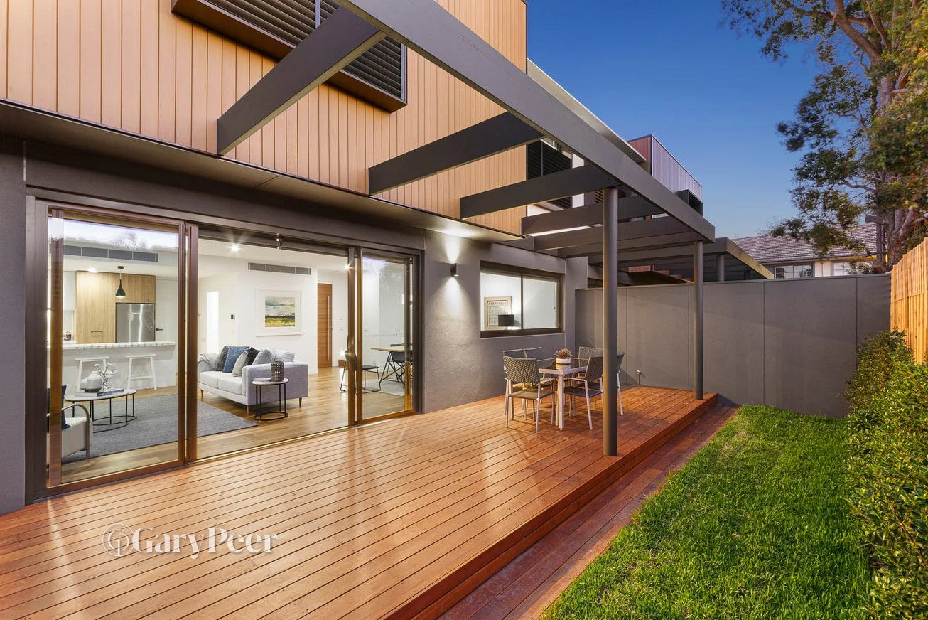 Modern apartment outdoor deck with wooden flooring, outdoor dining table with four chairs, and sliding glass doors leading into a living room.