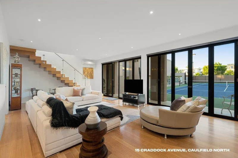 Modern living room with white sofas, a round chair, large sliding glass doors leading to a tennis court, wooden flooring, and a staircase