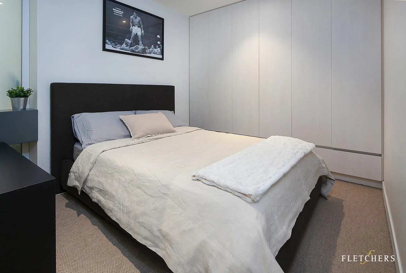 A neatly made bed with beige and gray bedding and pillows in a minimalist bedroom. There is a black and white photograph of a boxing match hanging on the wall above the bed. A white closet spans the wall to the right, and a small black side table wit
