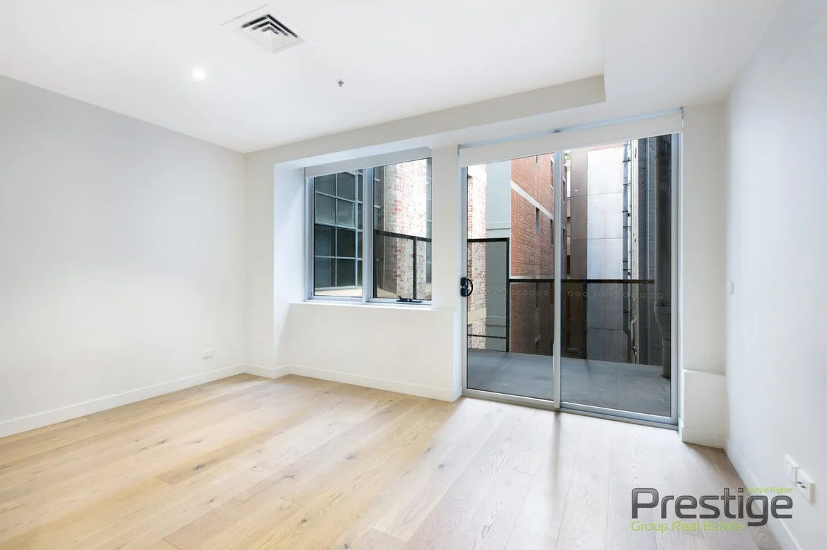 Empty apartment room with light wooden flooring, white walls, a window, and sliding glass door leading to a small balcony outside.