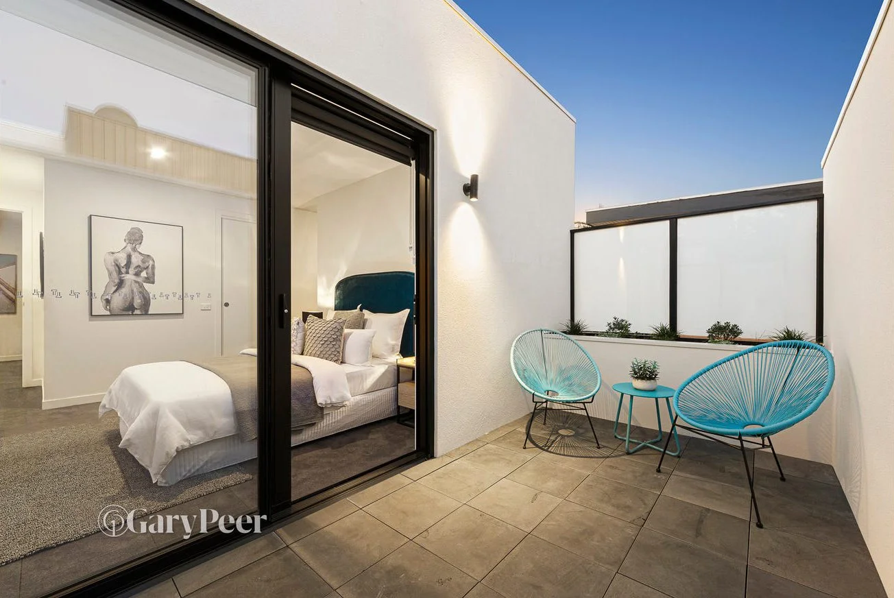 Modern bedroom with balcony featuring two blue wire chairs, a small round table with plants, and an interior space with a bed and art on the wall.