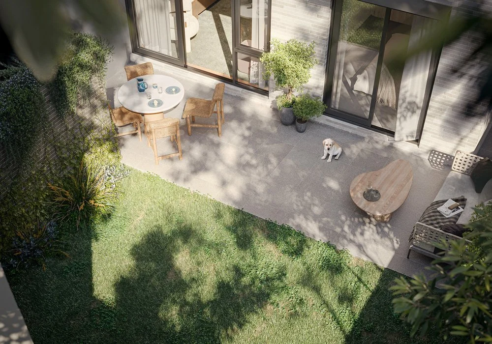A backyard patio with a white round table and three wooden chairs. A small dog sits on the tiled area near potted plants, a coffee table, and outdoor furniture. There is a grassy area with trees and shrubs, and a sliding glass door leading inside.
