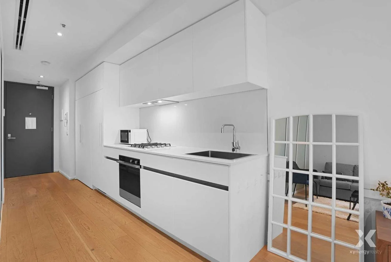 Modern kitchen with white cabinets, black countertop, microwave, stove, oven, and a sink with a faucet. A decorative mirror and living room with couch are visible through a windowed partition.