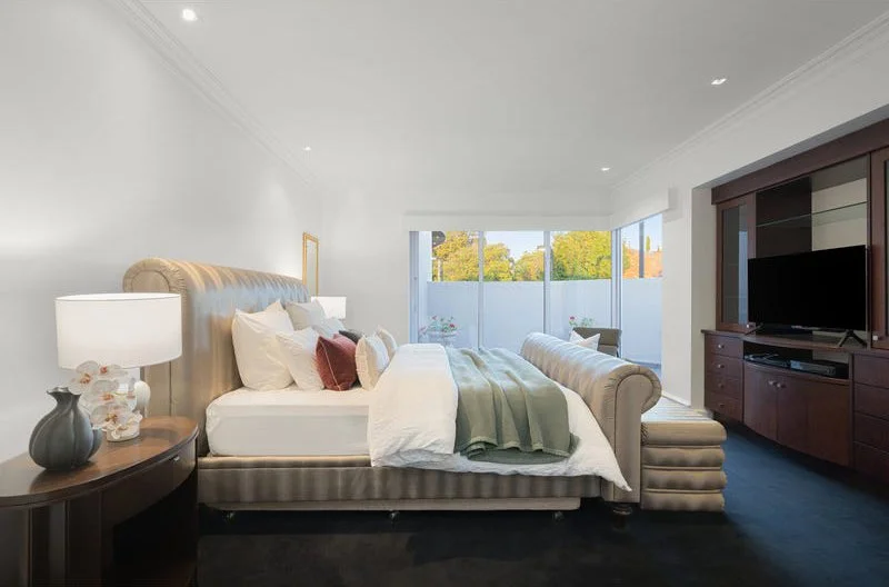 A modern bedroom with a large bed, nightstands, lamps, a window with a view of trees, and a television cabinet.