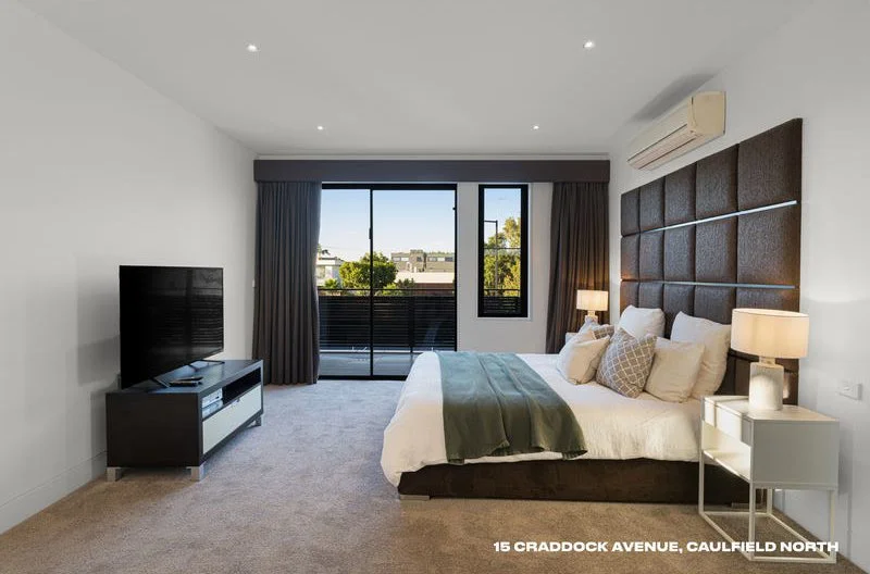 Modern bedroom with a large bed, pillows, bedside lamps, a TV on a stand, and a balcony view with buildings and trees outside.