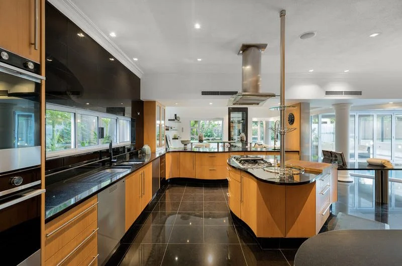 Modern kitchen with black flooring, wooden cabinets, a large island with a gas stove, stainless steel appliances, and large windows letting in natural light.