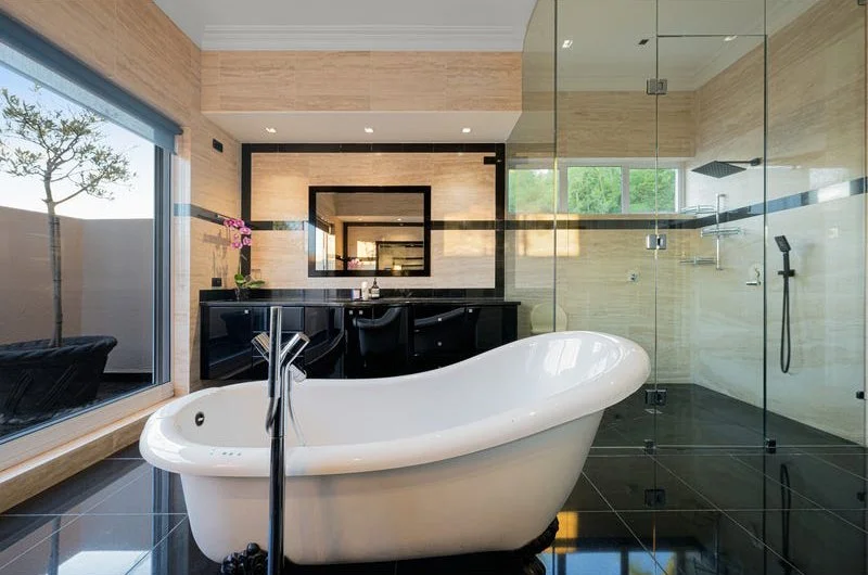 Modern bathroom with a freestanding bathtub, a large glass shower enclosure, a black vanity with a sink, and a large window with a view of trees and a patio.