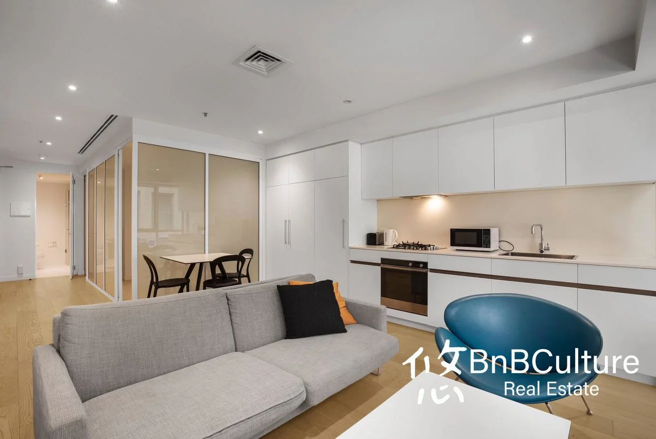 Modern living room and kitchen with grey sofa, blue chair, white cabinets, and wooden flooring.