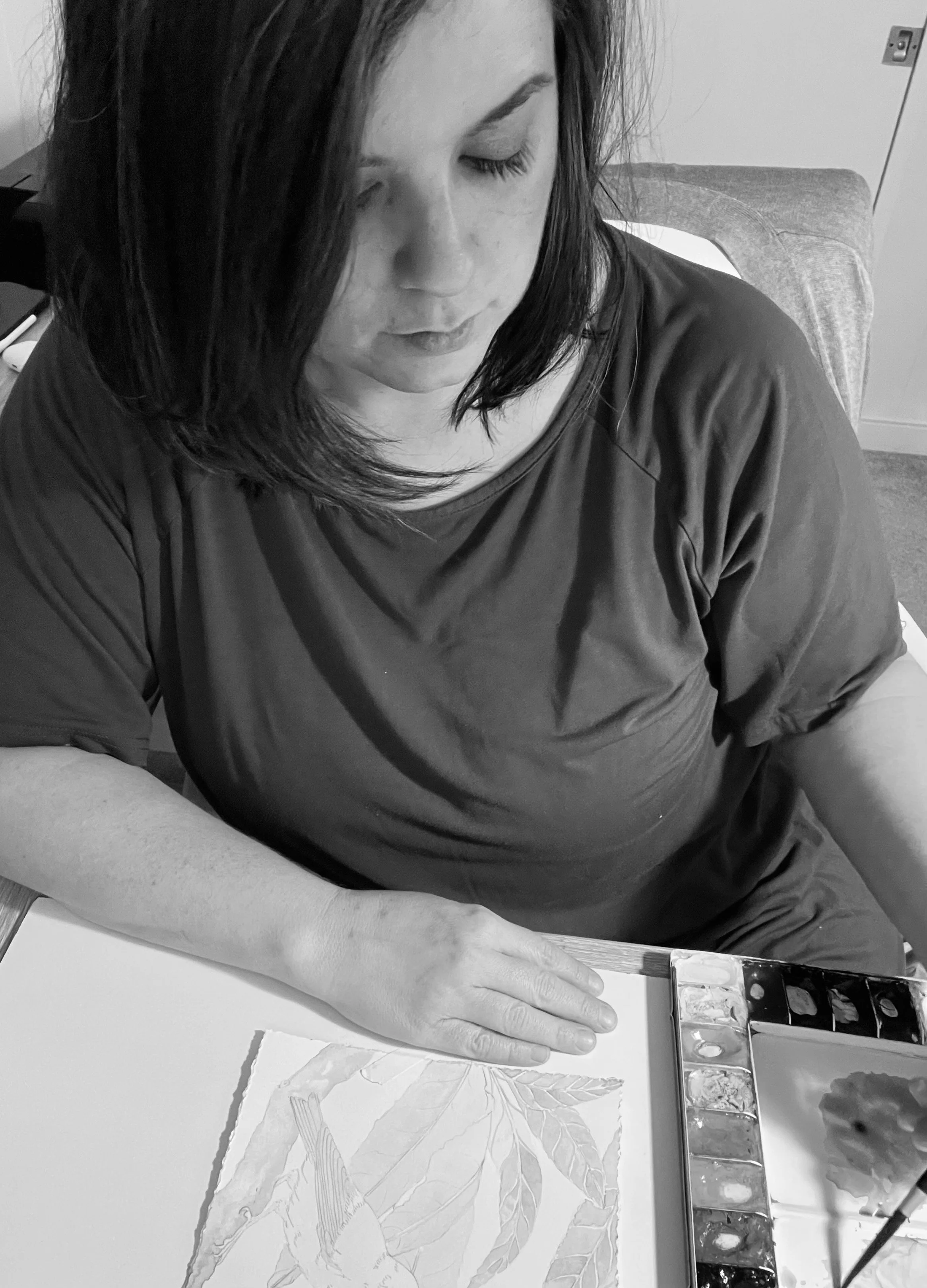 A woman with dark hair looking down at a watercolor painting of leaves and a branch on a table.