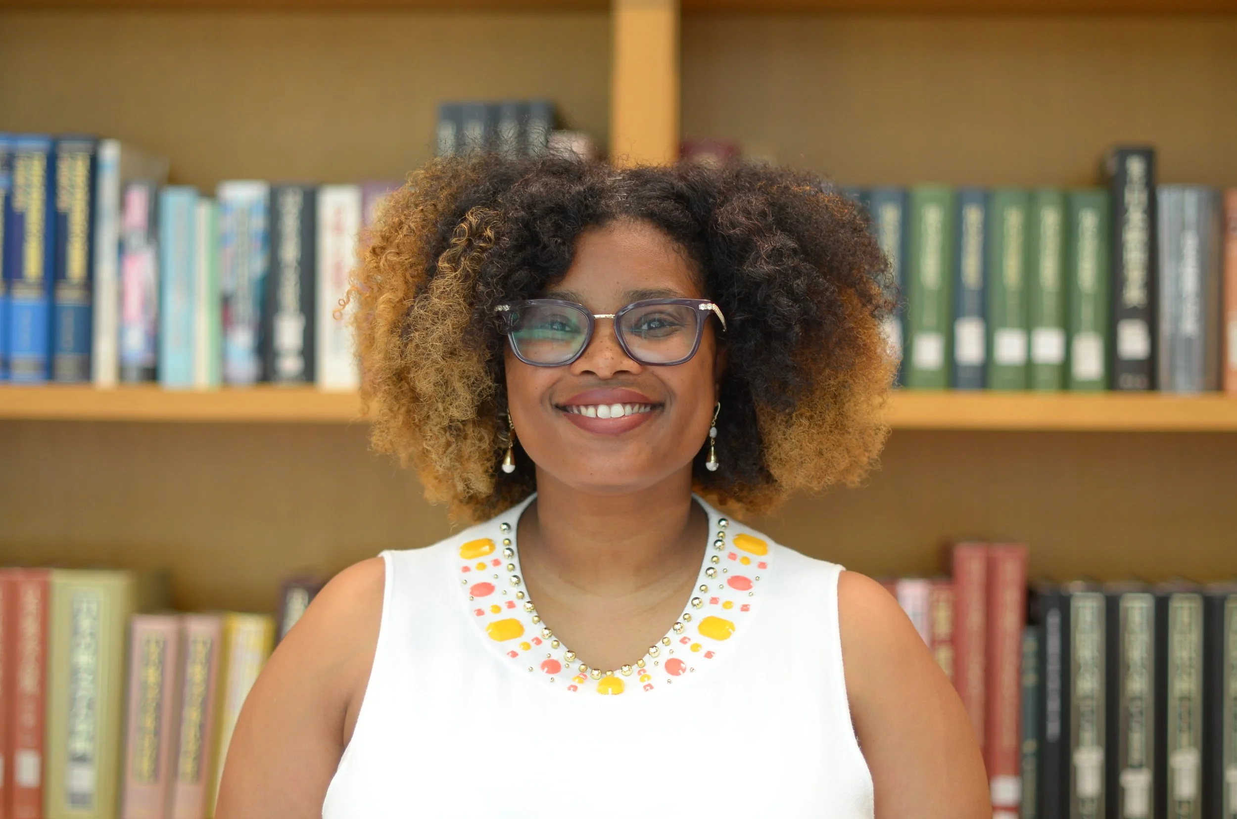 Image of Dr. Caprice Phillips smiling with a glasses on and a white blouse with a colorful neck. Standing in front of a bookcase.