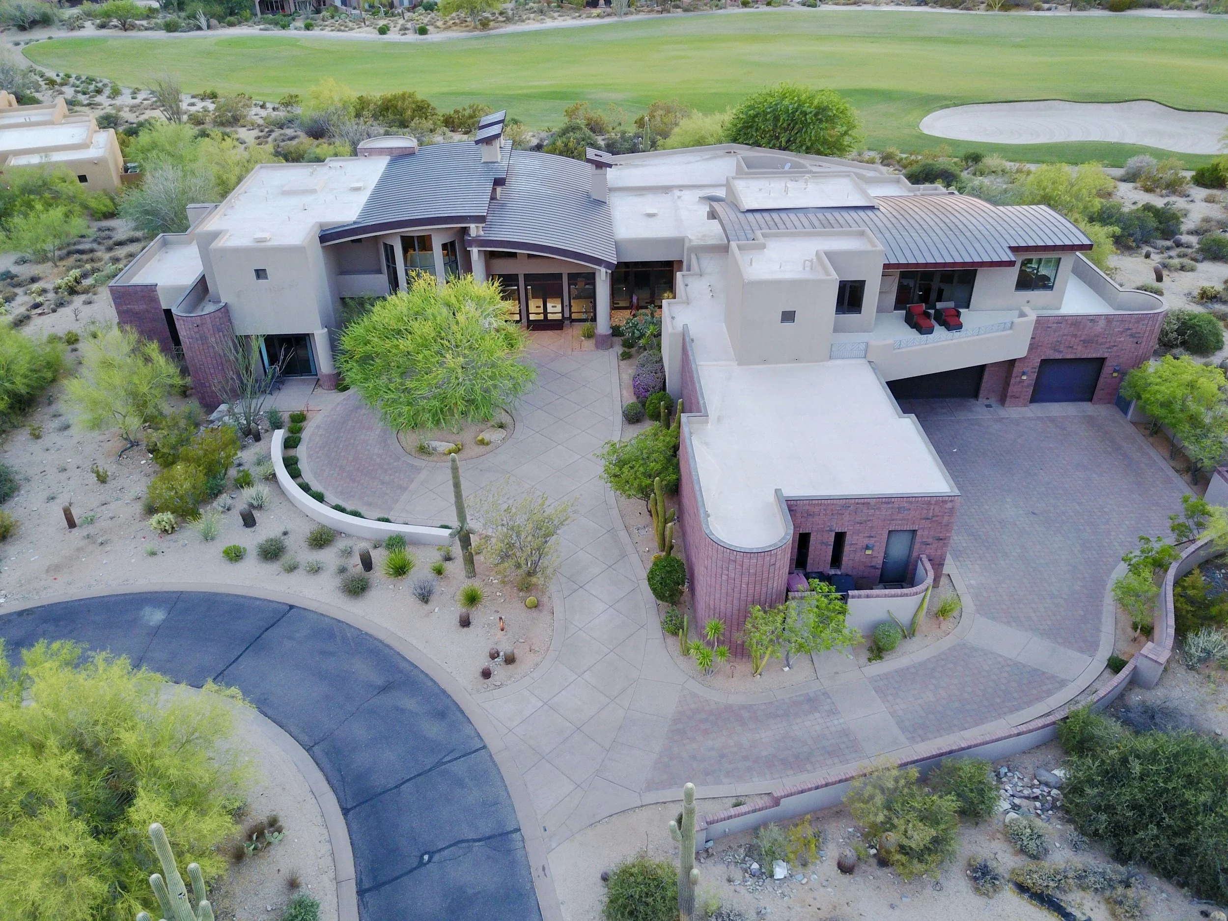 Aerial view of a modern house with flat and curved roofs, surrounded by desert landscaping, large trees, and a backyard patio, with a golf course in the background.