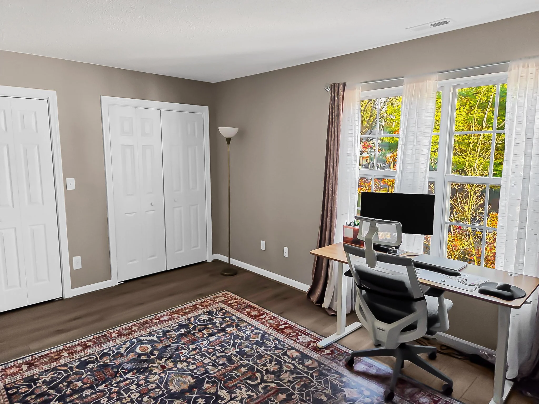 A home office with a white desk, black and gray office chair, large window with white curtains and a view of trees, beige walls, and a patterned area rug on the wooden floor.