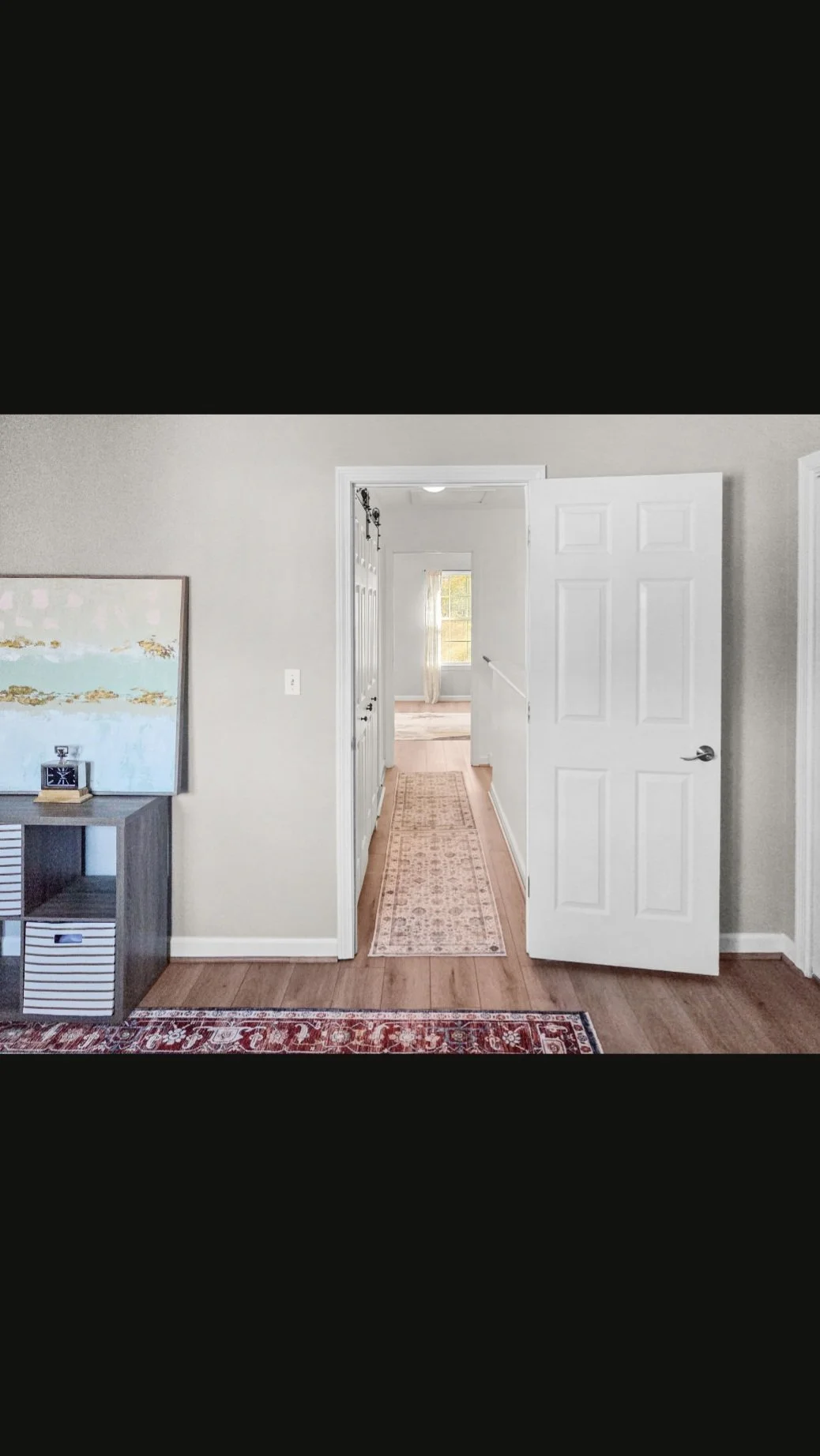 View of a hallway through an open white door in a living room, with a narrow runner rug on the hardwood floor, distant window with curtains, and light-colored walls.