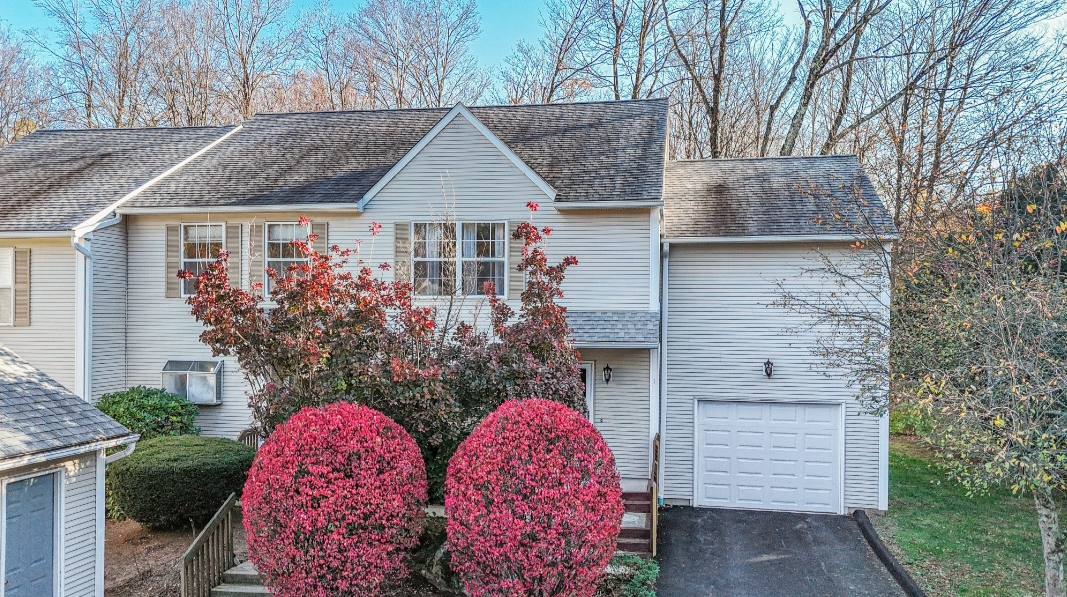 Front of a two-story house with white siding, a garage, and a small set of stairs leading to the front door. Two large pink bushes and a flowering tree are in the front yard, under a partly cloudy sky.