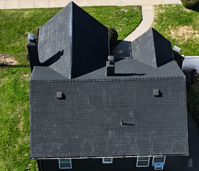 Aerial view of a house with a gray shingle roof, chimneys, and surrounding green lawn with a sidewalk.