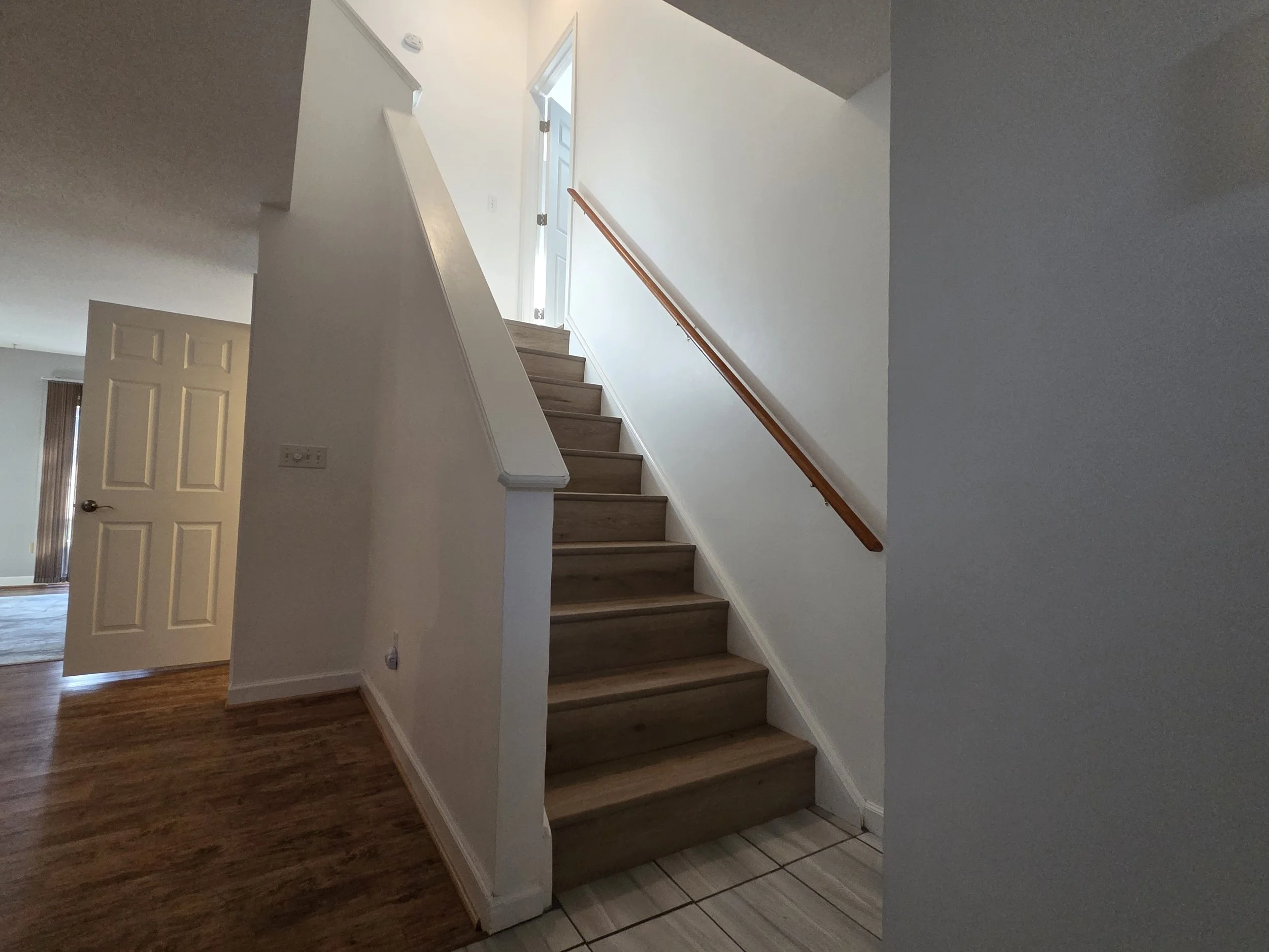 A staircase with wooden steps and a wooden handrail, located next to a white wall in a home interior. A door and some parts of a room are visible in the background.