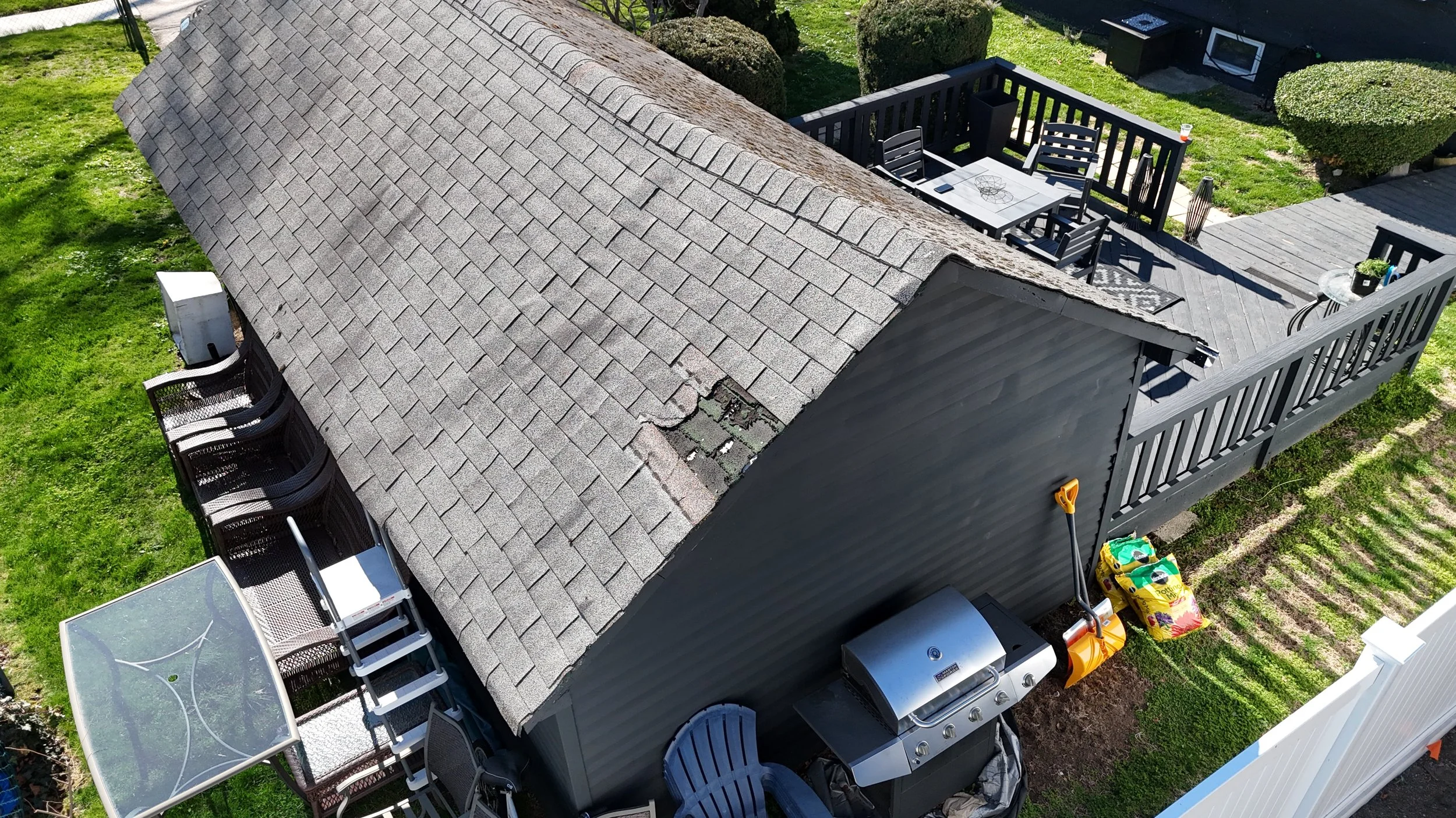 Aerial view of a backyard featuring a shed with a shingled roof, a wooden deck with outdoor furniture, a small table, a barbecue grill, and various potted plants, with grassy areas and bushes surrounding the space.