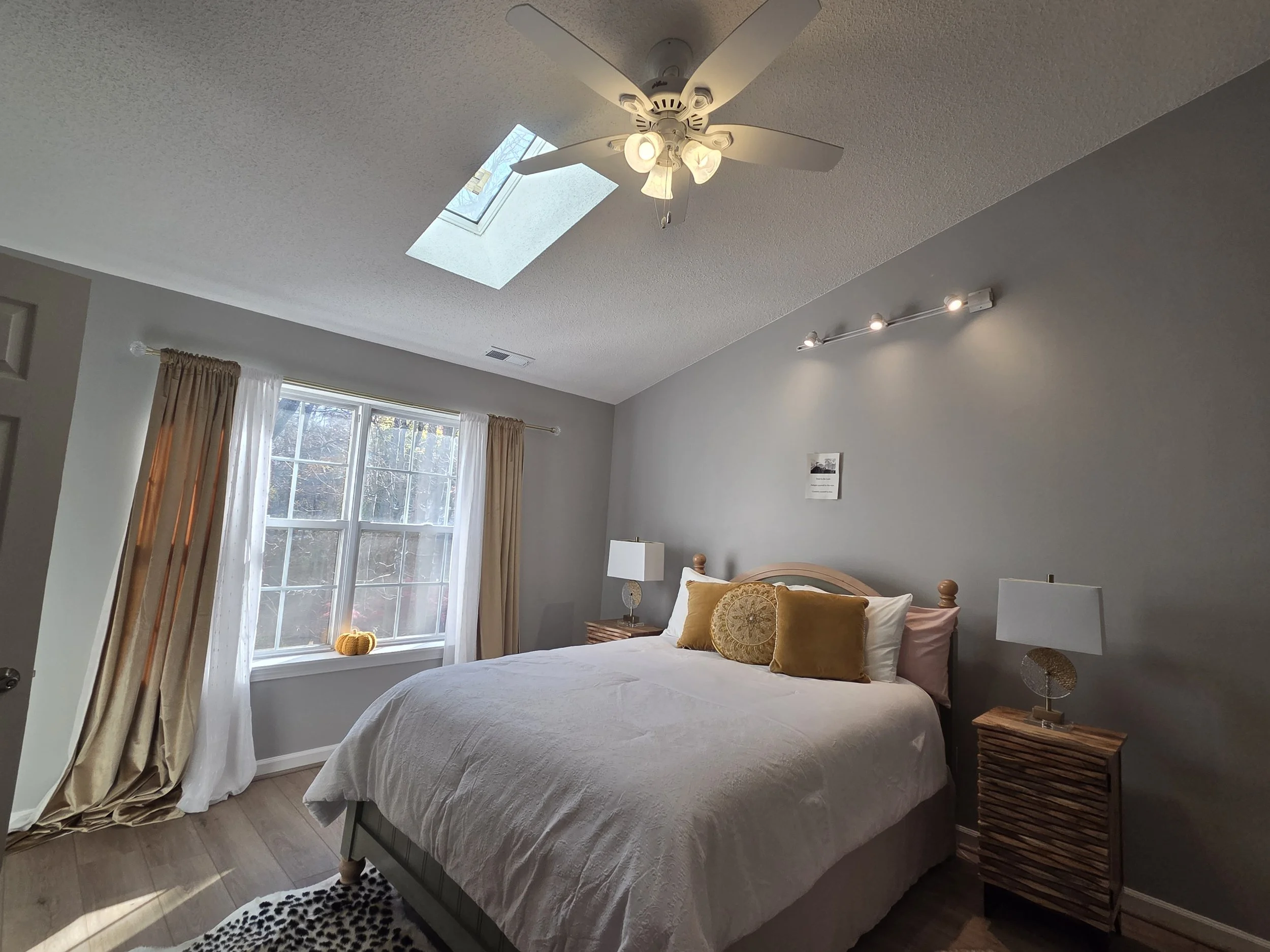 Bedrooms with large window, beige and white bedding, lamps, and a ceiling fan with skylight.
