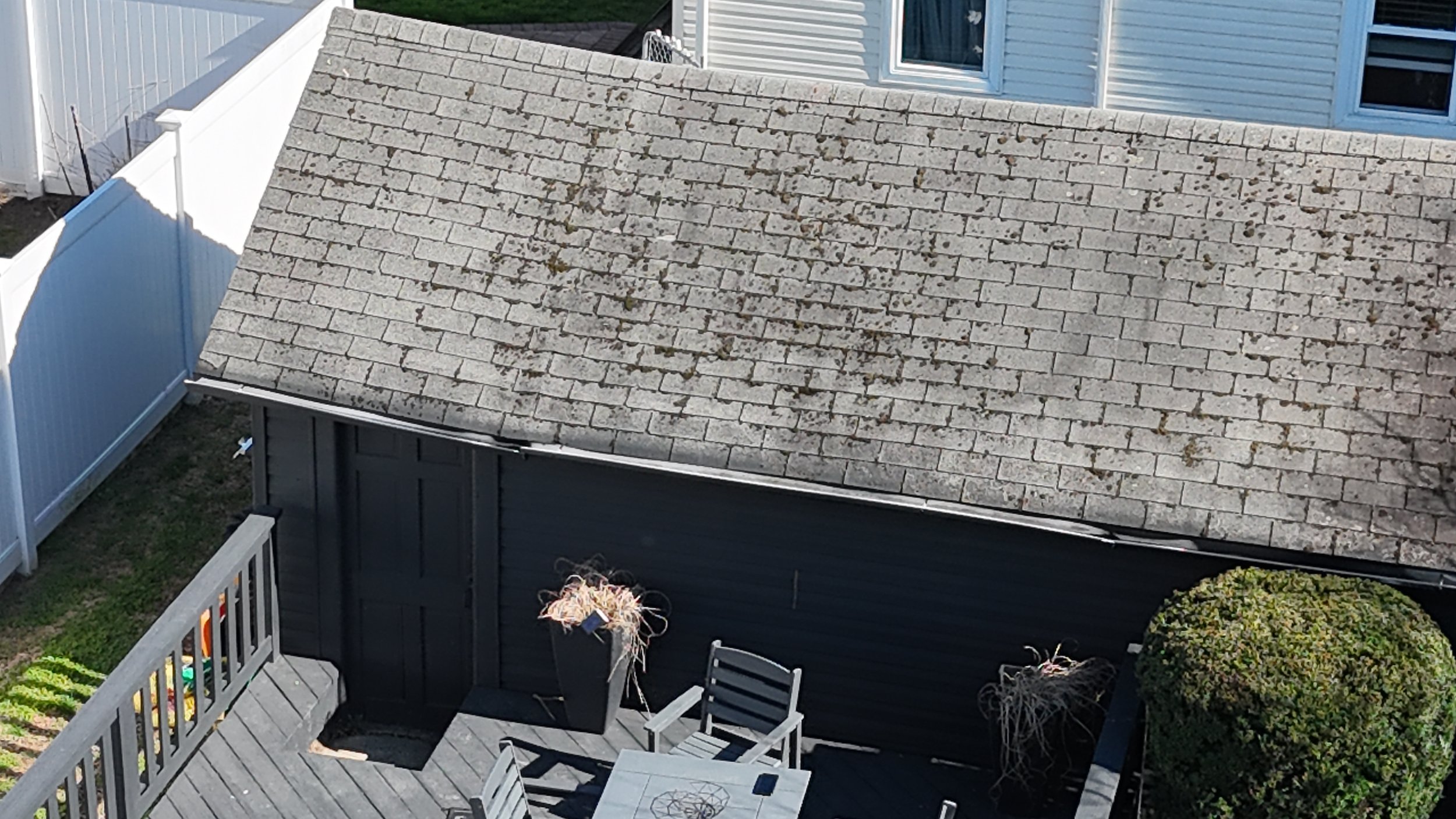 An aerial view of a backyard patio area featuring a gray wooden deck, outdoor table with chairs, a small adjacent shed, and a fence surrounding the space.