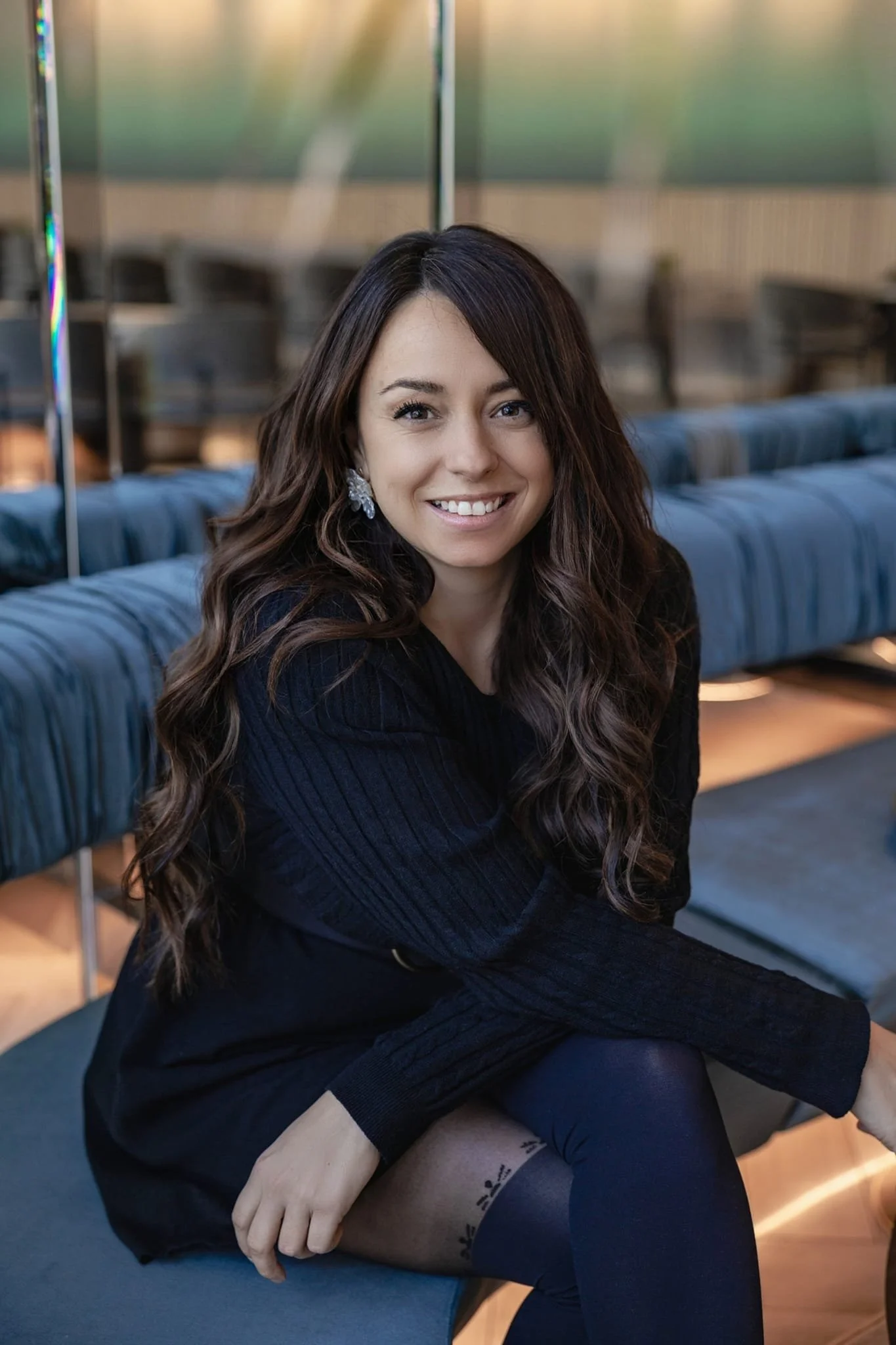 Jeune femme aux longs cheveux bruns, souriante, assise sur un sofa bleu, portant une robe noire et des collants noirs avec un motif floral en bas.