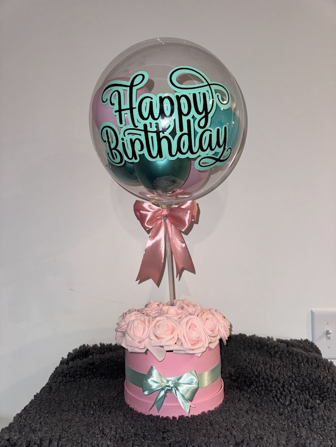 Stuffed bubble balloon full of mini-balloons on a stand inside a flower box with soft pink rose heads and Vinyl wording.