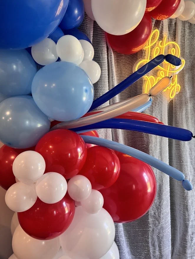 Split Organic Balloon Garland with red, white and blue balloons, foil star balloons, balloon pearl strand, white curtain, and LED Let's Party sign.