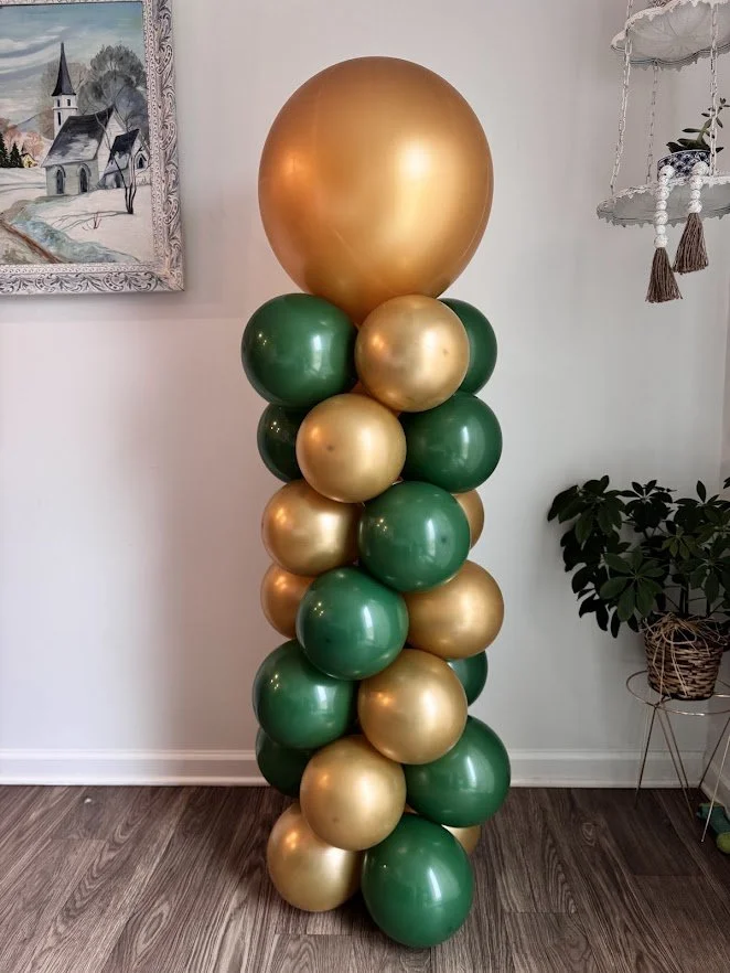 A tall column of green and gold balloons arranged vertically, topped with a large gold balloon, standing in a room with white walls, a painting on the wall, and a potted plant nearby.