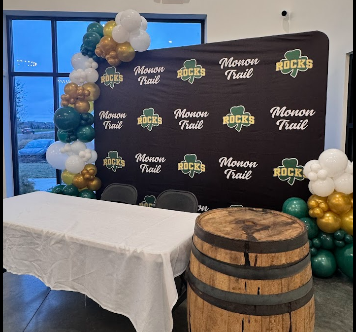 Event setup with Monon Trail and Rocks branding, decorated with green, white, and gold balloons, featuring a table covered with a white cloth and a barrel in front.