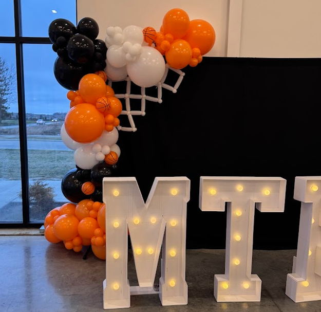 Decorative marquee letter 'M', with a balloon arch of black, white, and orange balloons, some styled as basketballs, against a black backdrop near a window.