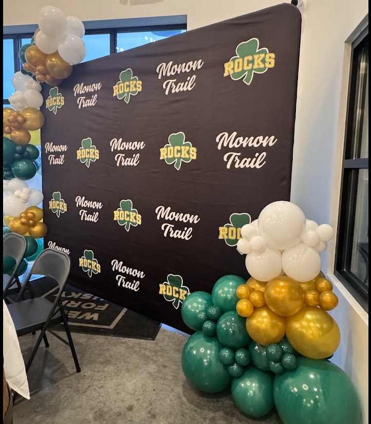 Decorative backdrop with logos reading "Monon Trail" and "Monon Rocks," surrounded by green, gold, and white balloons arranged in clusters.