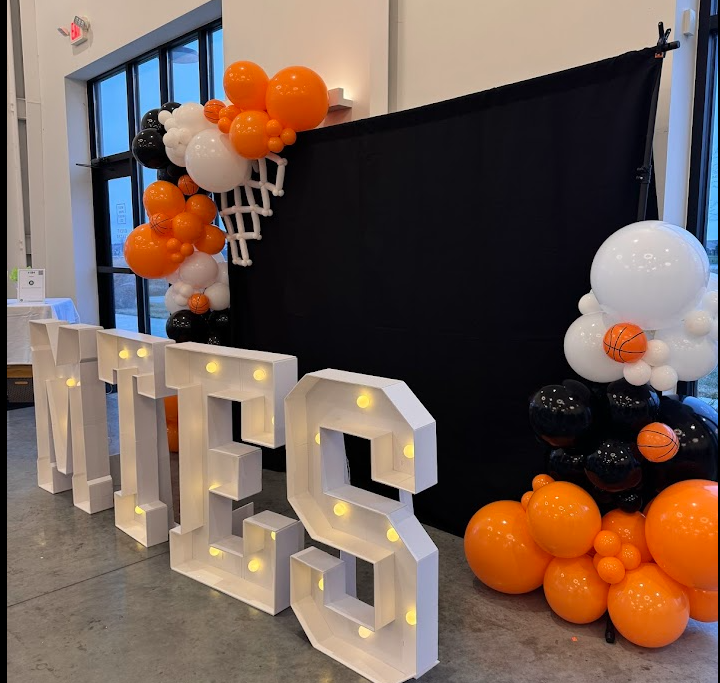 Decorative display for a Halloween themed event with large illuminated white letters spelling 'BOO', surrounded by orange, black, and white balloons, some of which feature basketball designs, and a black backdrop.