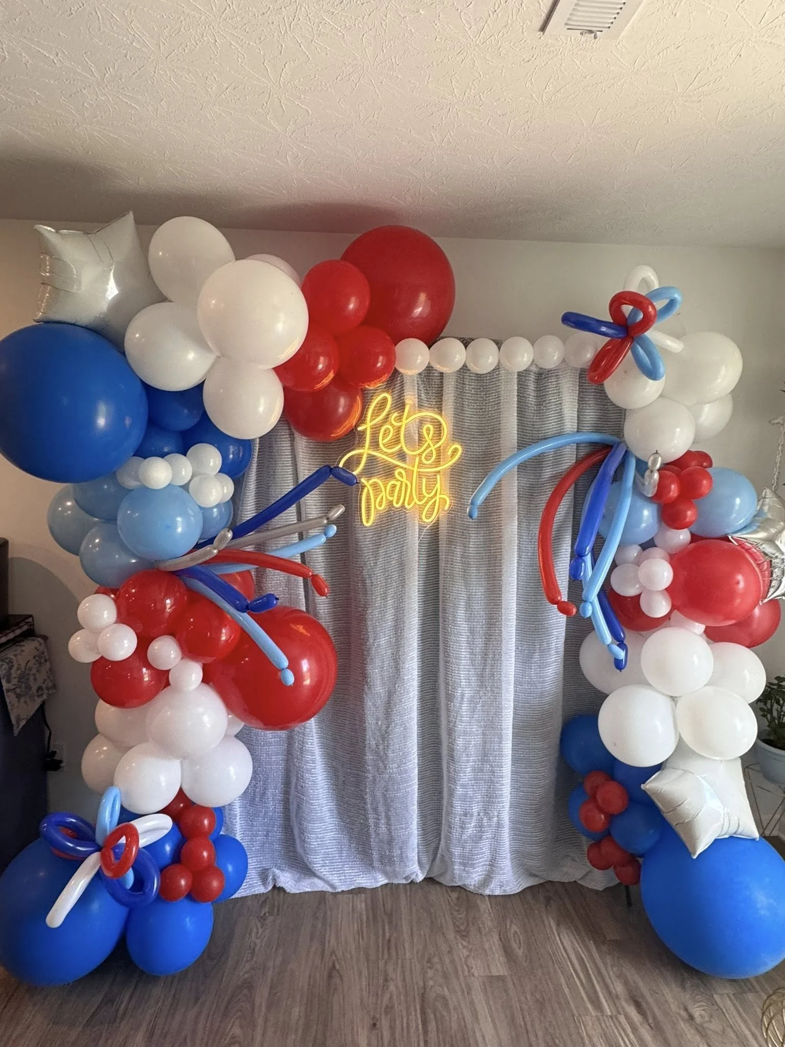 Split Organic Balloon Garland with red, white and blue balloons, foil star balloons, balloon pearl strand, white curtain, and LED Let's Party sign.