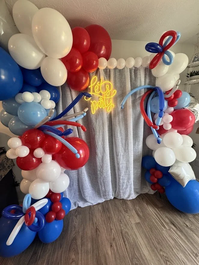 Decorative balloon arch in red, white, and blue colors with a neon sign that says 'Lets Party' and a gray curtain backdrop.