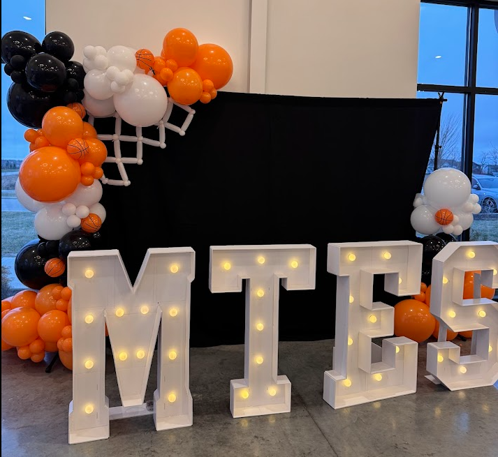 Decorative setup with illuminated marquee letters spelling 'MTES' in front of a black backdrop, adorned with black, white, orange, and basketball-themed balloons clustered on both sides. Large venue area for staging. 