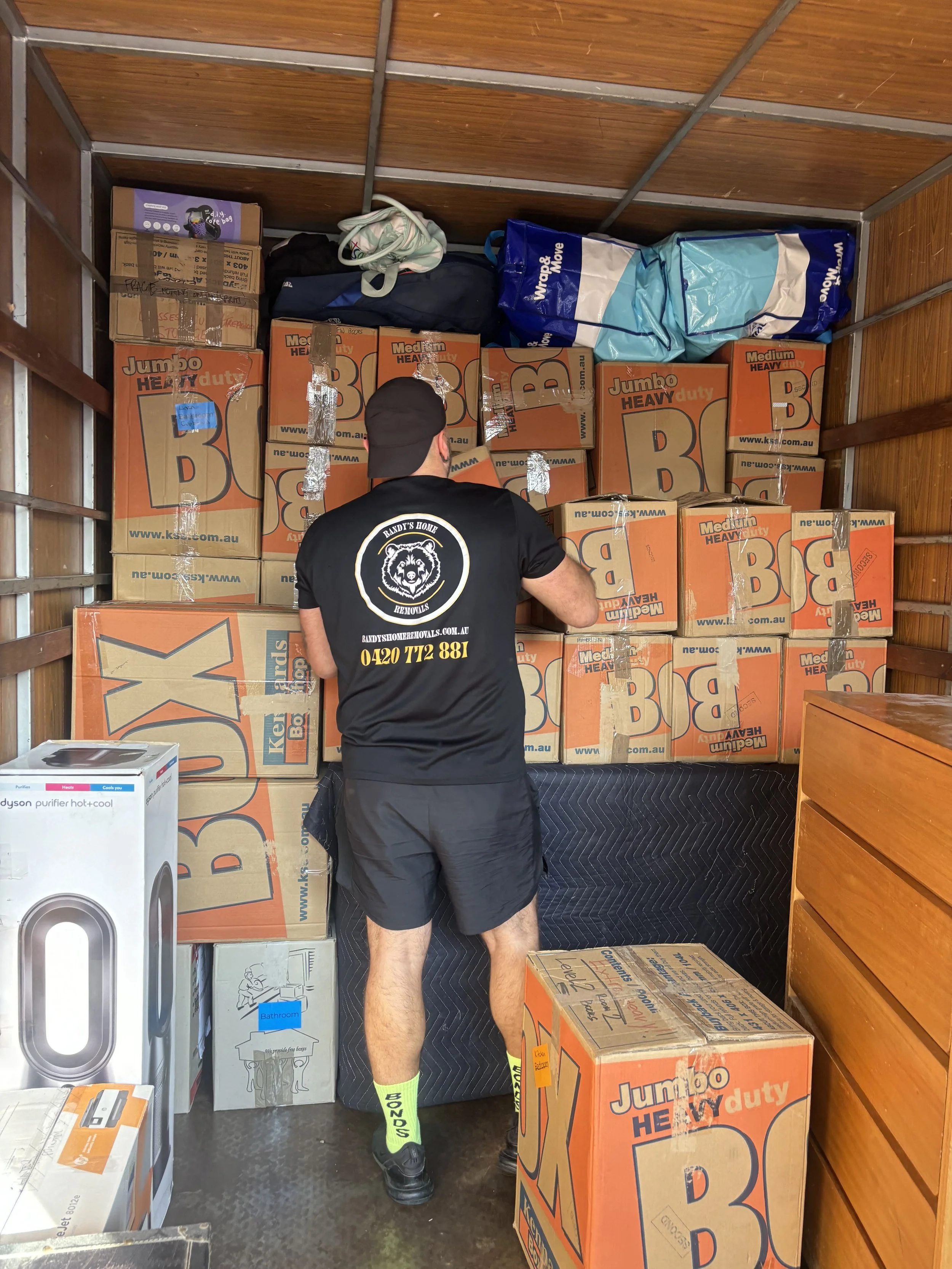 A person standing in a small storage space filled with packed cardboard boxes labeled "Jumbo Heavy Duty B" and some other items like a Dyson air purifier and bags, with their back facing the camera.