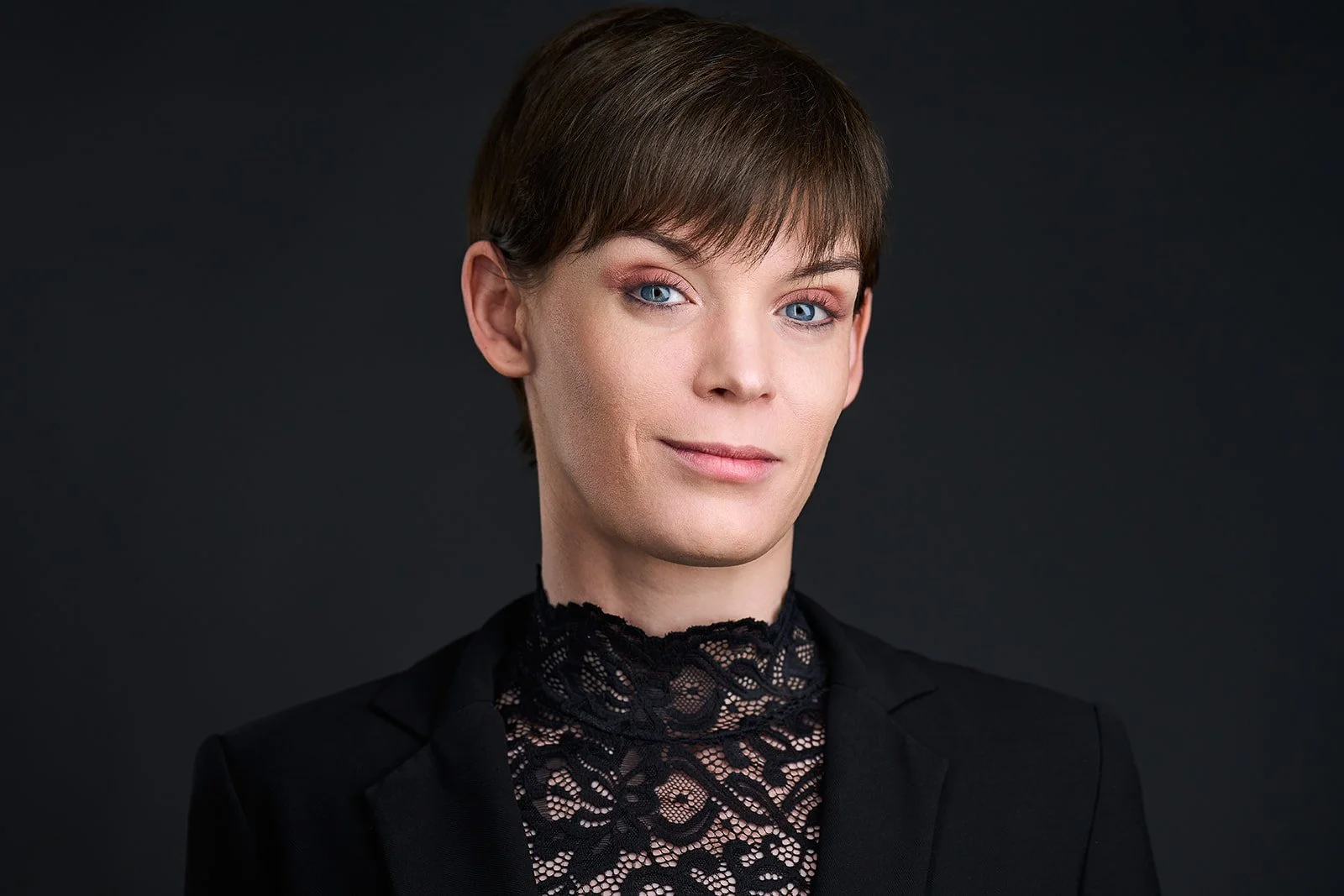 Portrait of a woman with short brown hair, blue eyes, and light skin, wearing a black blazer and a lace black top, against a dark background.