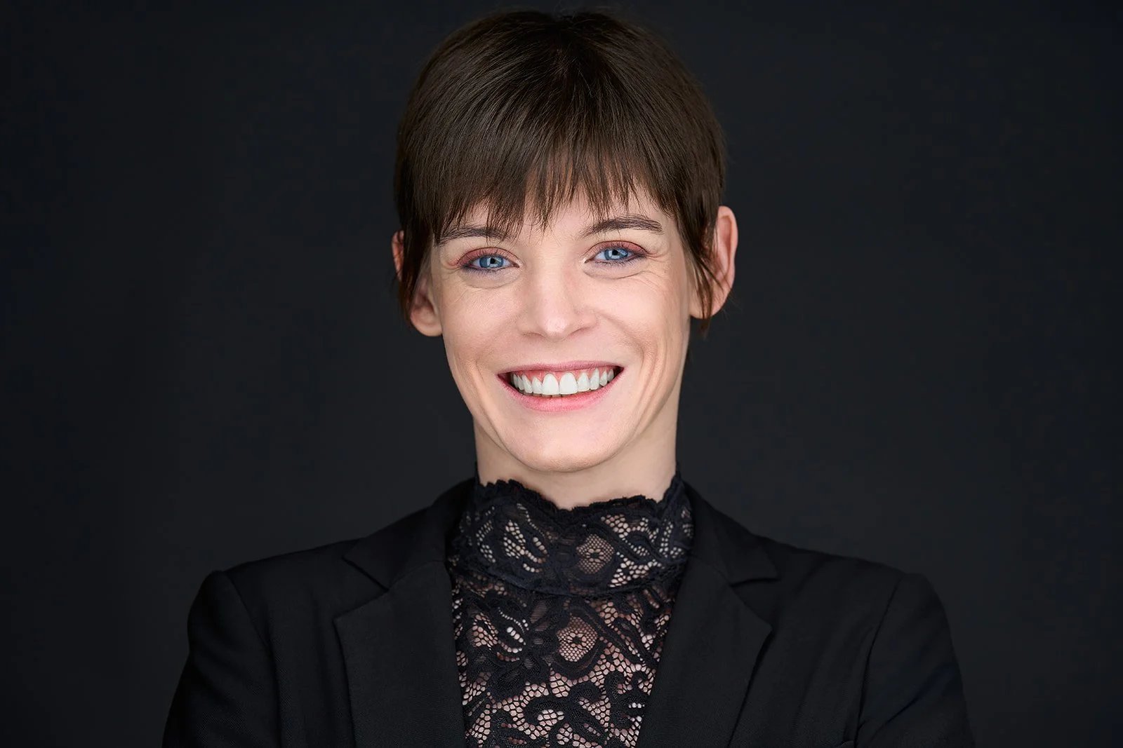 Portrait of a smiling woman with short brown hair, blue eyes, wearing a black lace top and blazer, against a dark background.