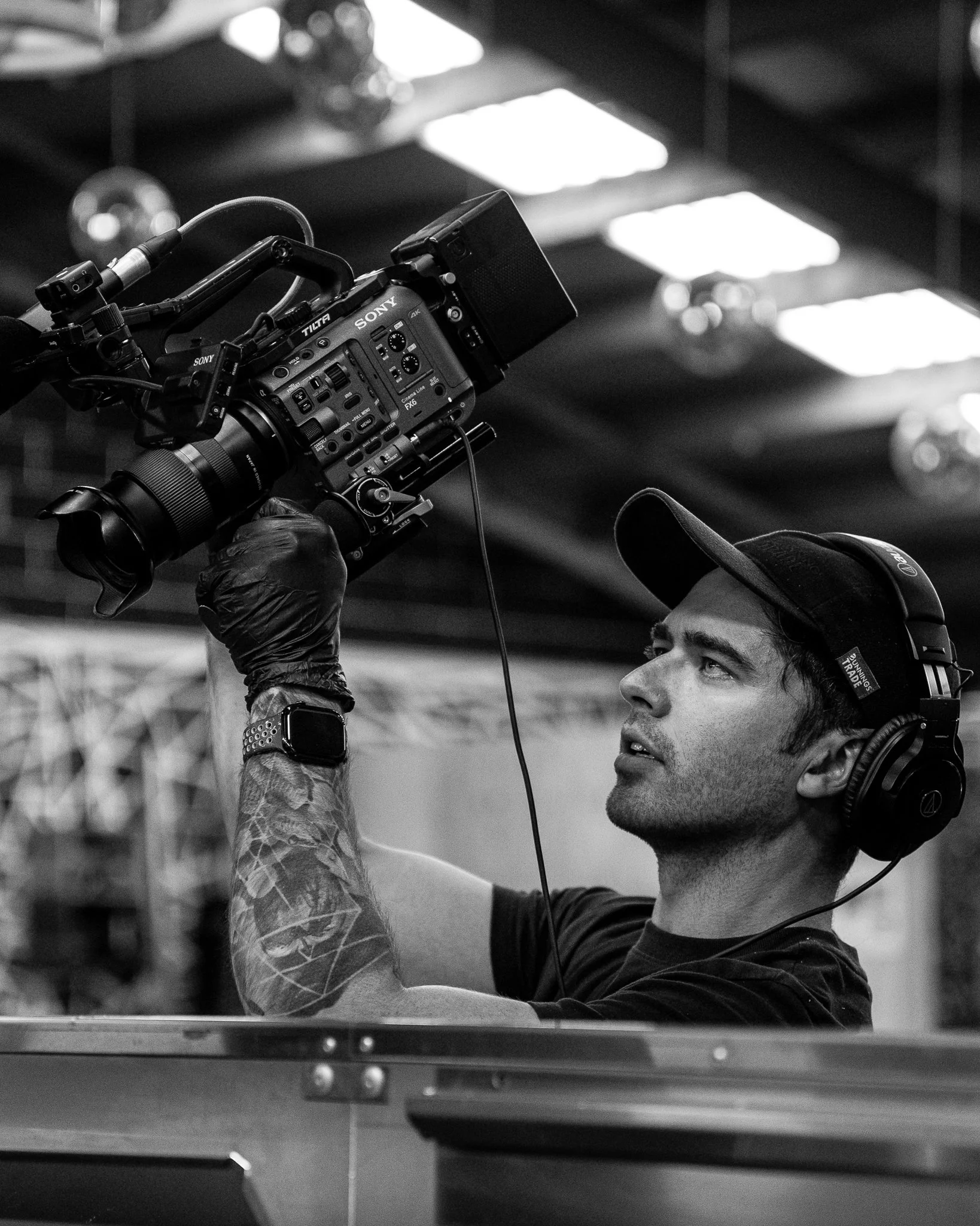 A man wearing headphones and a cap operating a professional Sony video camera indoors.
