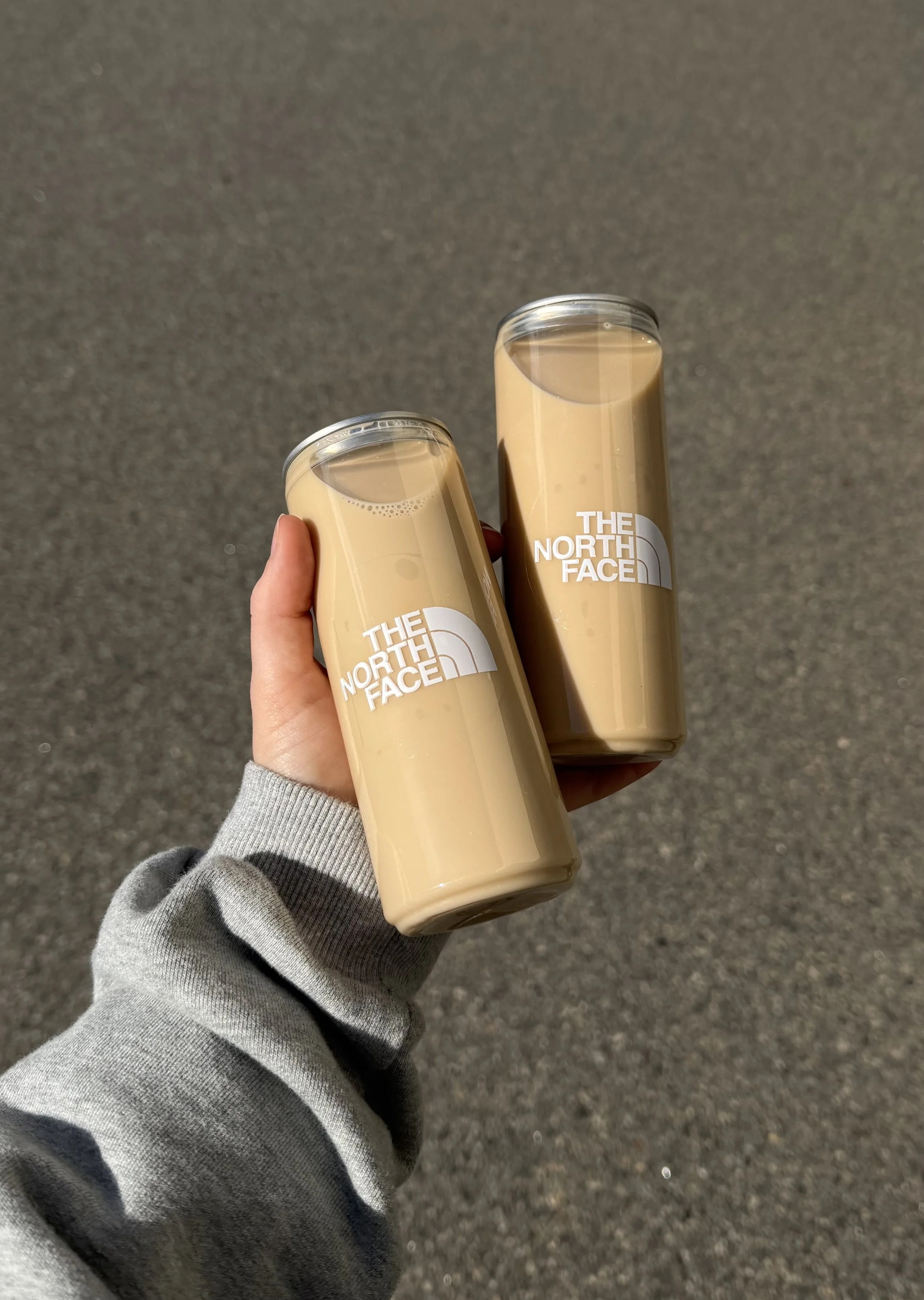 Two cold beverages in clear plastic cups with 'The North Face' logo, held by a person wearing a gray sweatshirt, against a gray sidewalk background.