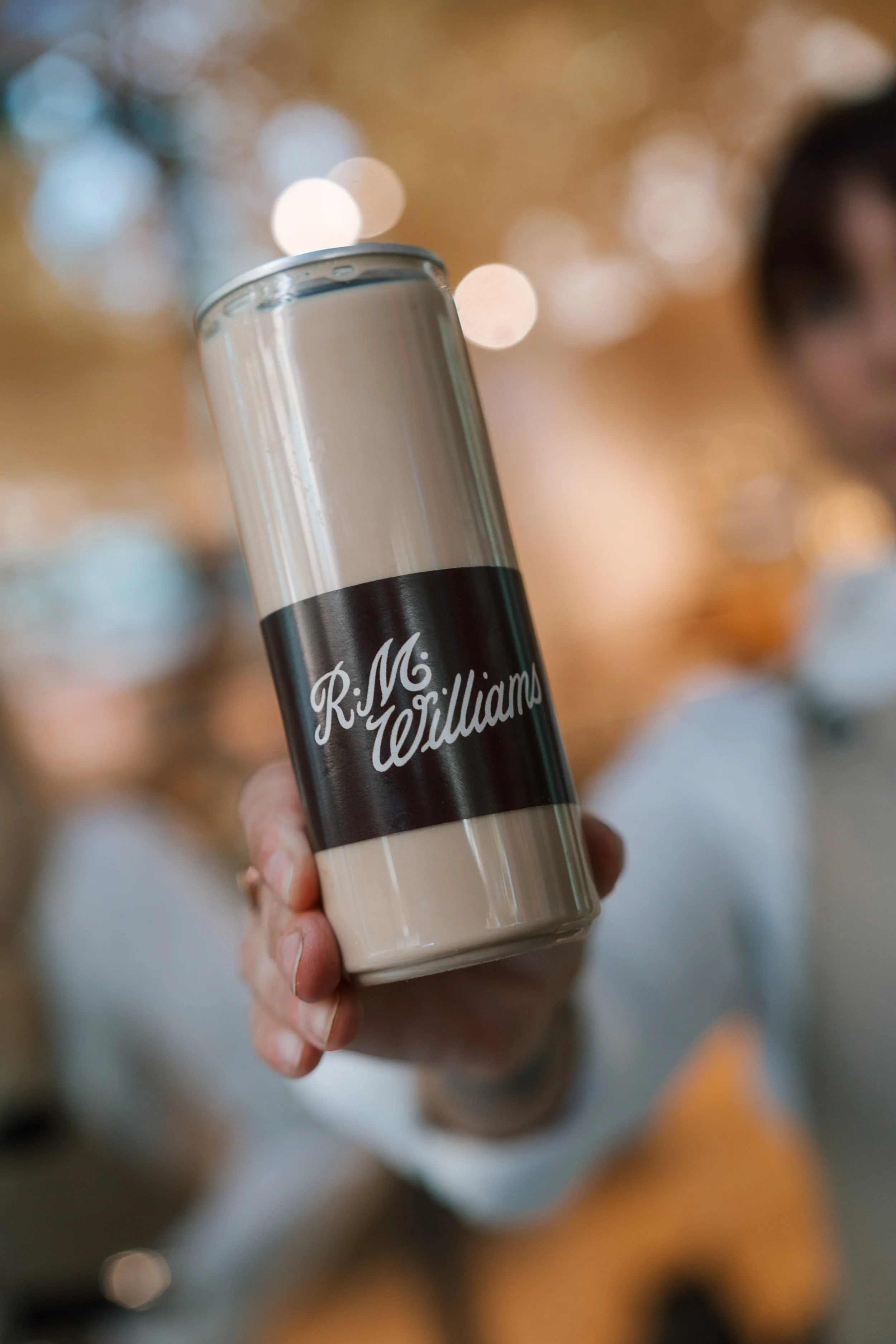 Close-up of a hand holding a beverage can with the text 'Ri Miliam' on it, blurred background with warm bokeh lights.