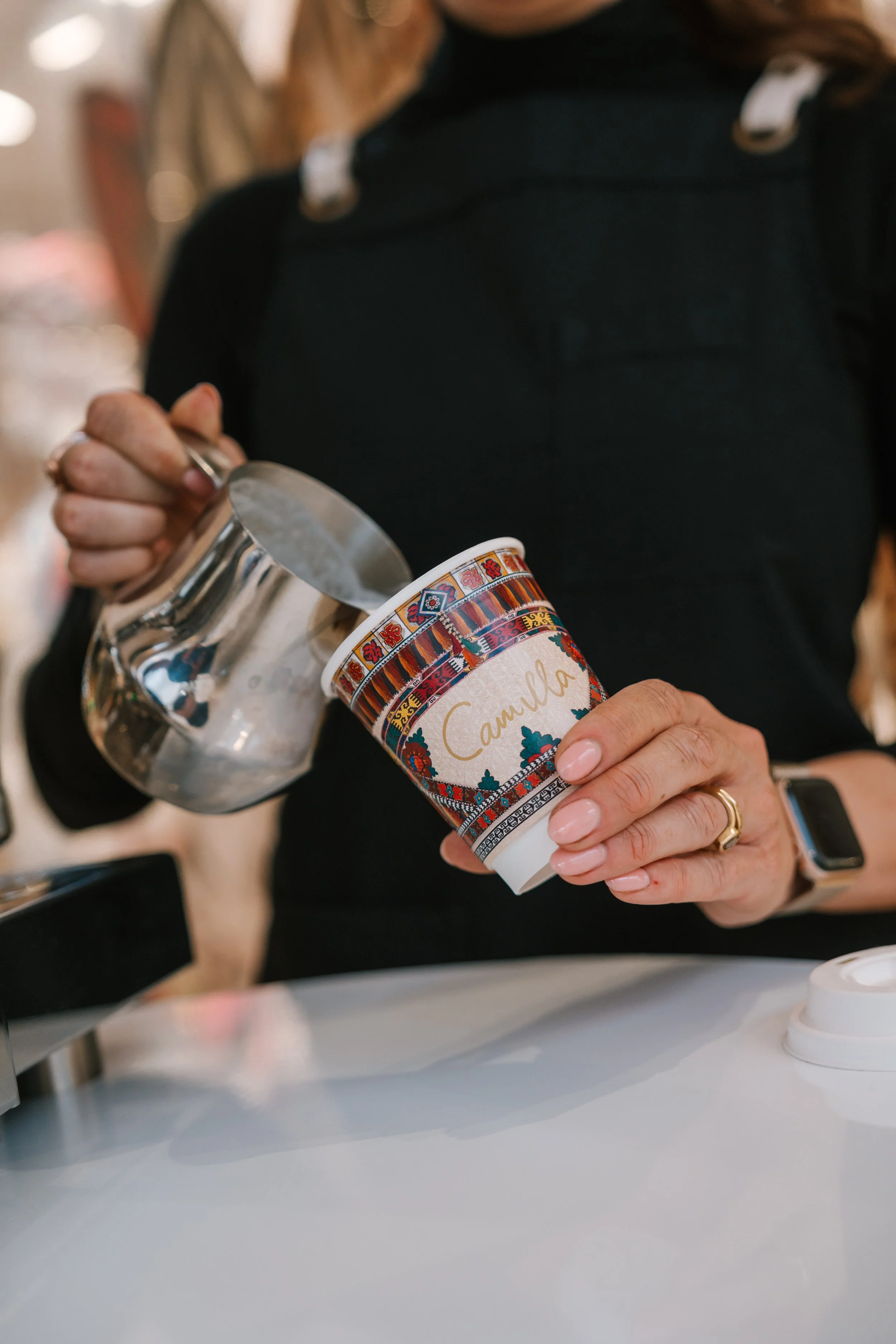 Person pouring water from a glass into a disposable coffee cup with a colorful design and the words 'Camulla' on it.