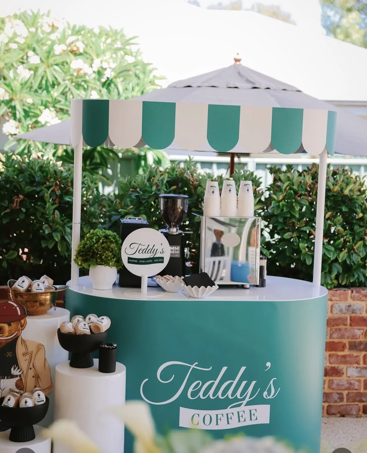 Coffee cart with a teal canopy and sign, featuring cups, a coffee machine, and decorative plants outdoors.
