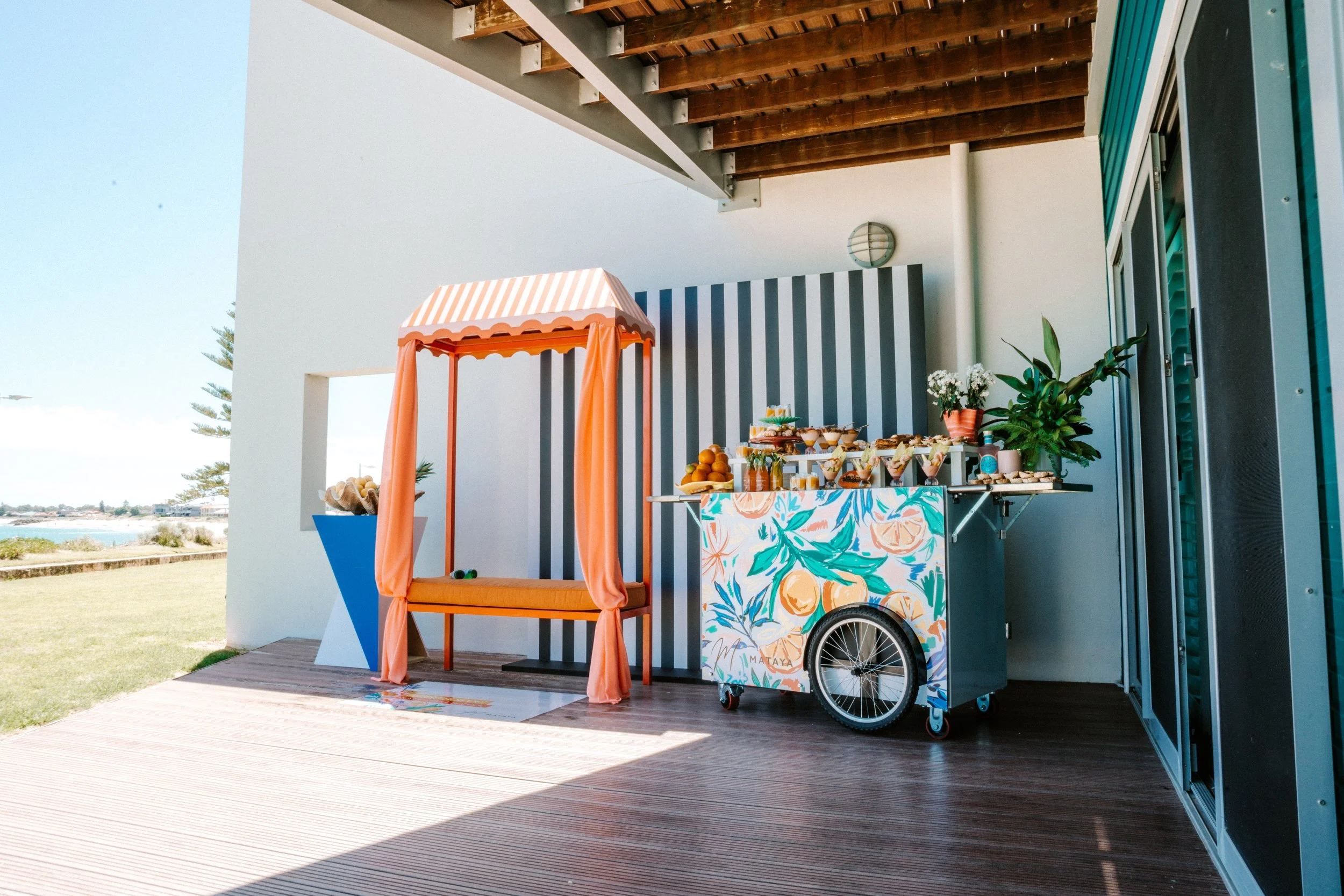 An outdoor setting with a small snack cart decorated with a colorful floral design, a striped awning, and a stool with orange fabric. The snack cart is stocked with fruits, drinks, and snacks. There is a table near the cart with more food and drinks.