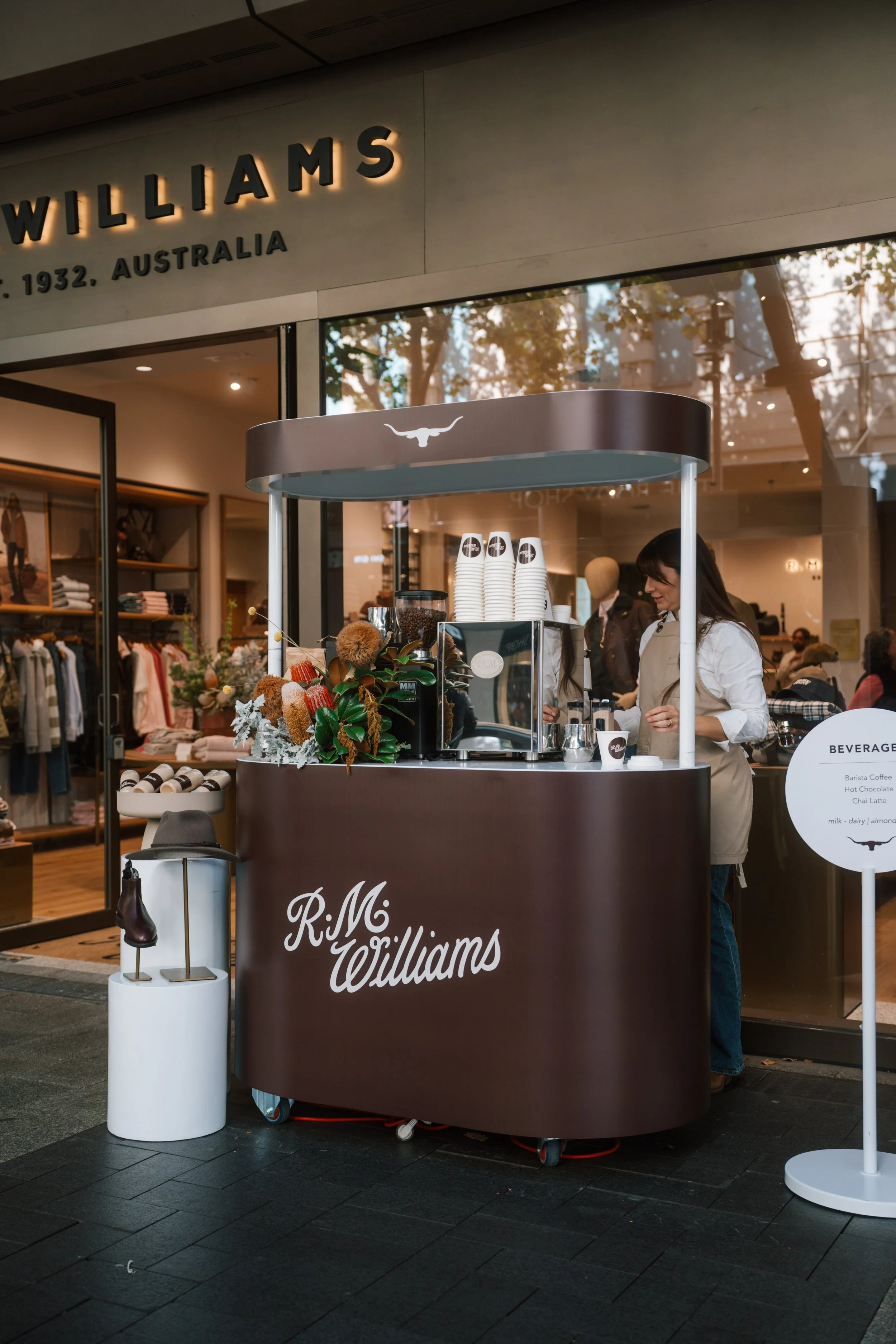 A food cart with the name R.M. Williams, serving beverages like hot chocolate, chai latte, and milk, with a woman operating it inside a shopping mall, Williams store in the background.