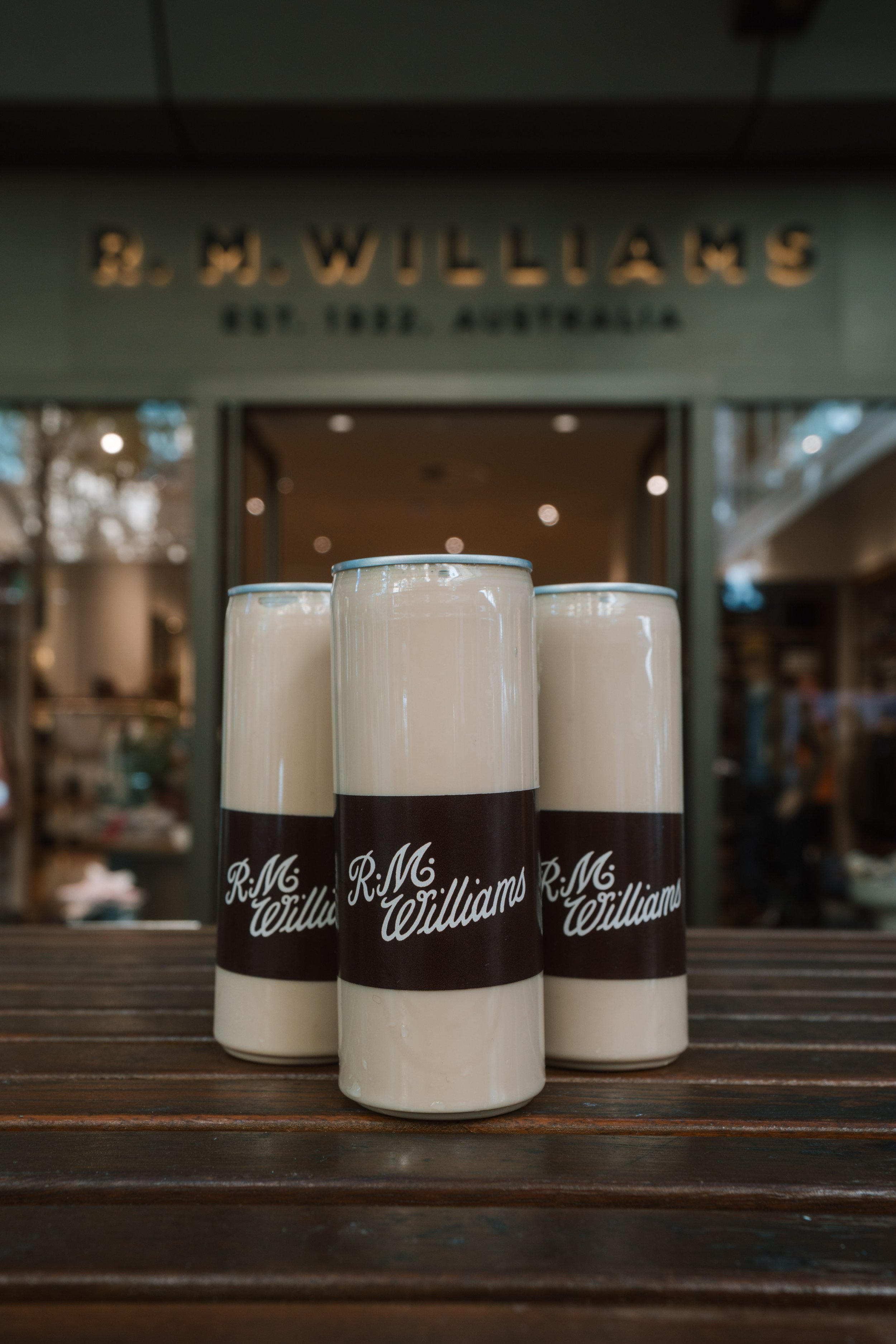 Four cans of R.M. Williams branding placed on a wooden surface with a store or shop background.