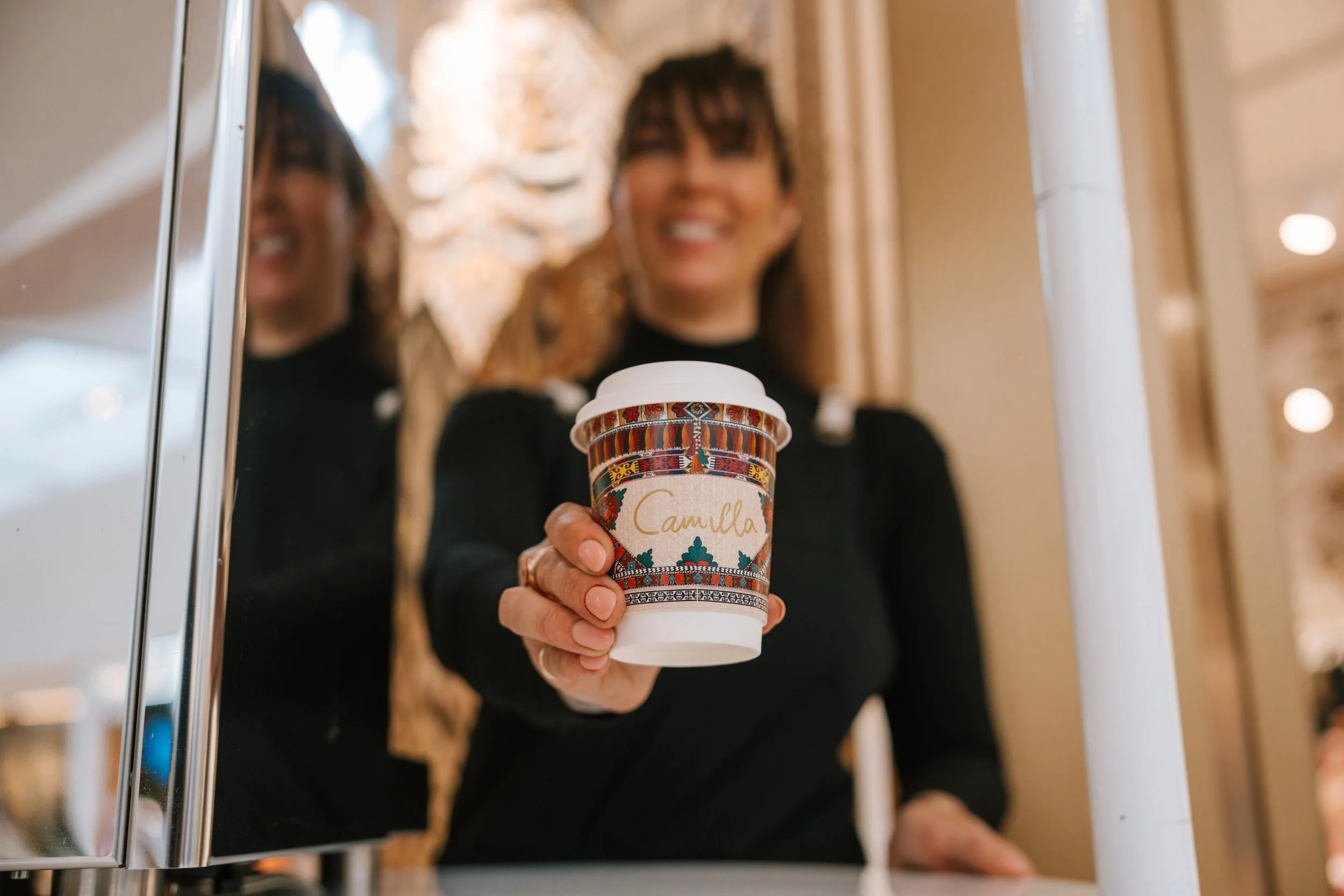Person holding a decorated travel coffee tumbler with the name 'Camilla' printed on it, seen in a reflection in the mirror with warm interior lighting.