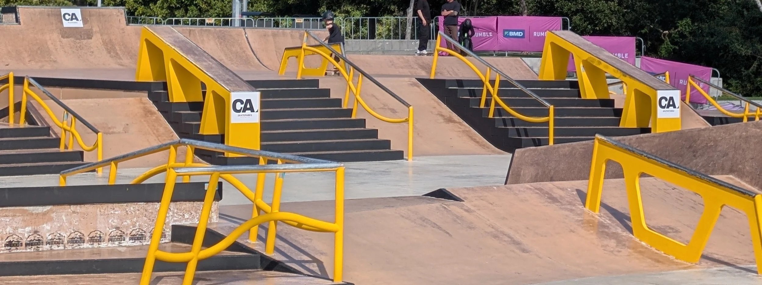 Skate park with multiple ramps, stairs, and yellow safety rails, and a few people in the background near purple banners.